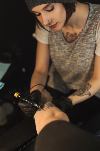 Tatuador de mujer concentrada haciendo tatuaje con máquina de tatuaje en el brazo masculino, vista superior. Profesión femenina inusual, espacio de copia