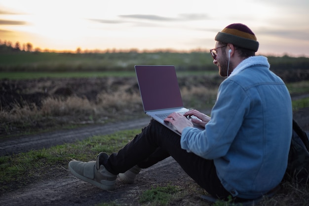 Tausendjähriger Hipster-Mann, der am Laptop mit Kopfhörern im Feld bei Sonnenuntergang arbeitet