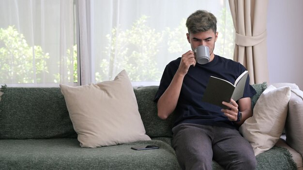 Tausendjähriger Mann trinkt heißen Kaffee und liest Buch auf dem Sofa zu Hause