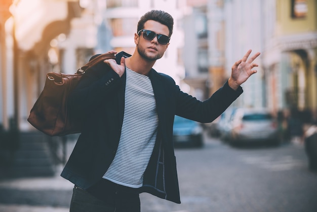 Táxi! Jovem bonito em traje esporte fino carregando bolsa no ombro e gesticulando enquanto pega carona na rua