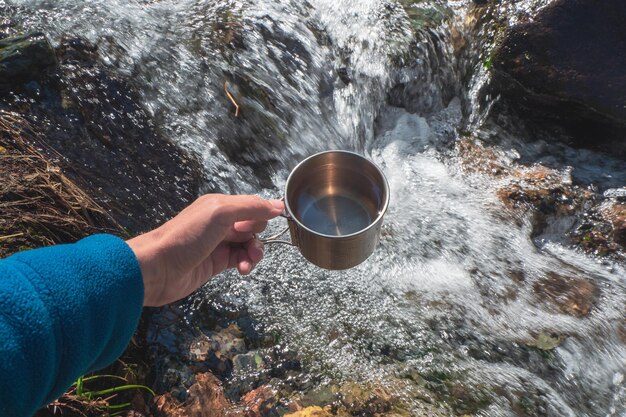 Taza con agua pura de glaciar de un arroyo de montaña. Mano sosteniendo una taza de excursionistas con agua potable limpia.