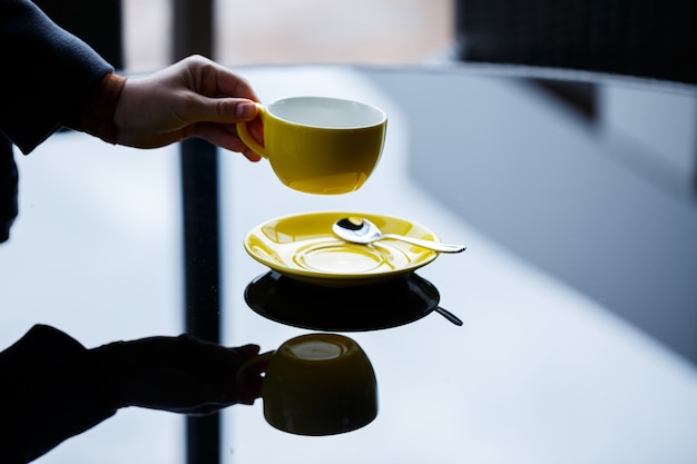 Taza amarilla para café o té con un platillo en manos de una niña en el fondo de una mesa de cristal.