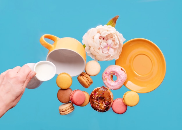 Foto taza amarilla vacía, donas, macarrones, peonía y mano sujetando la jarra de crema de leche volando sobre un fondo azul.