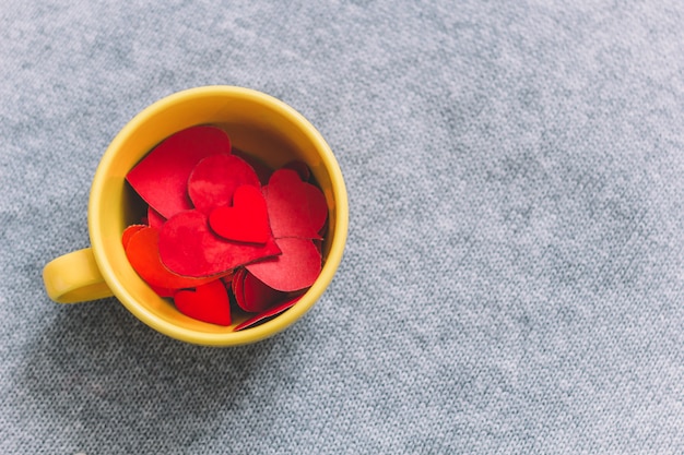 La taza amarilla de la vista superior llena de corazones rojos de papel en gris hizo punto el espacio de la copia del fondo