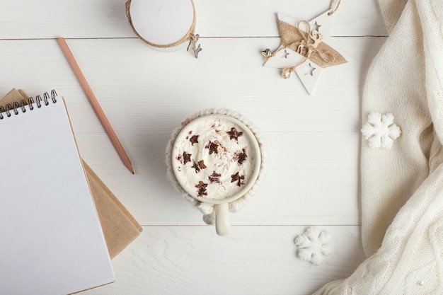 Una taza de bebida caliente de invierno, con crema batida y polvo con un asterisco, un cuaderno, copos de nieve blancos y una bufanda tejida sobre una mesa de madera.
