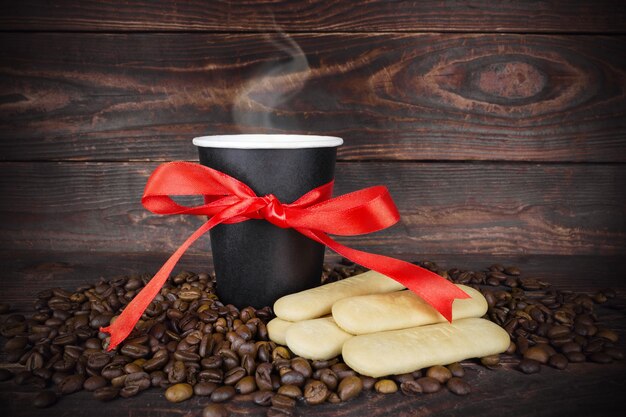 Taza de bebida para llevar desechable de papel negro con vapor arriba, con lazo hecho de cinta roja de regalo, granos de café esparcidos y galletas sobre fondo de tabla de madera. Café como regalo