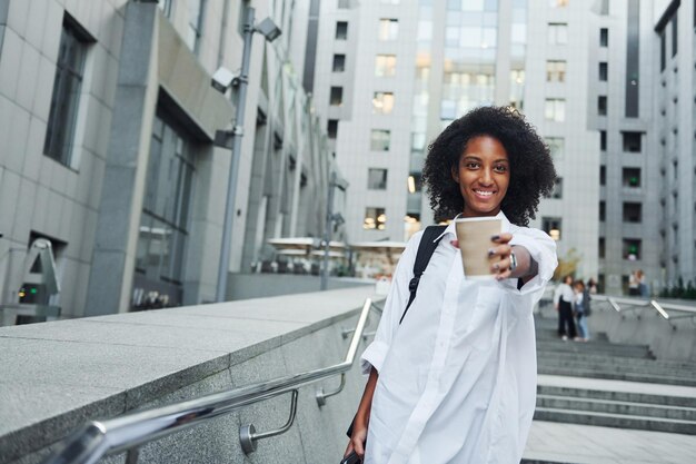 Con una taza de bebida, una mujer afroamericana vestida con buena ropa está al aire libre en la ciudad durante el día