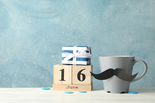 Taza con bigote decorativo, caja de regalo y calendario de madera sobre mesa blanca sobre fondo azul, espacio para texto