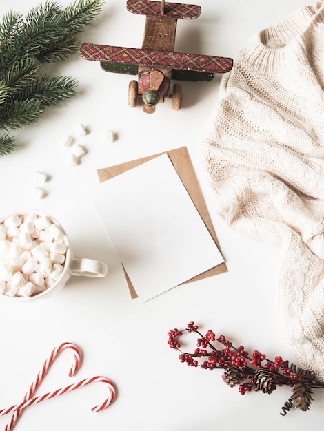 Taza blanca con bebida caliente y malvaviscos, tarjeta de papel para carta, sobre, cuadros de punto y decoración navideña