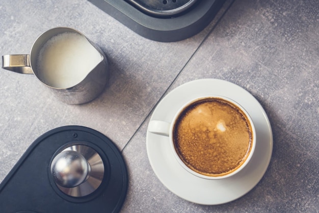 Foto taza blanca con café caliente recién hecho y leche en la mesa cerca de la jarra y la manipulación de la máquina de espresso