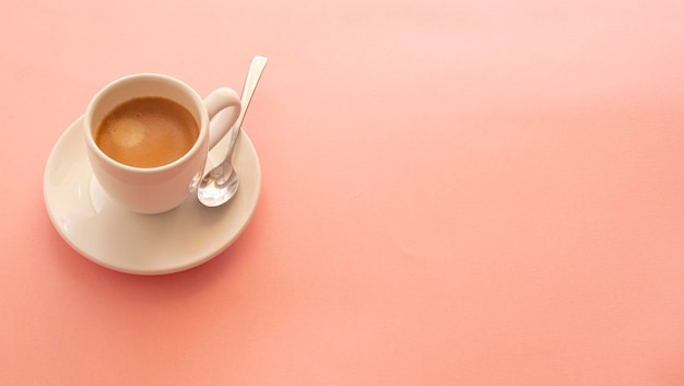 Taza blanca con café y una cuchara de metal brillante sobre un fondo rosa claro