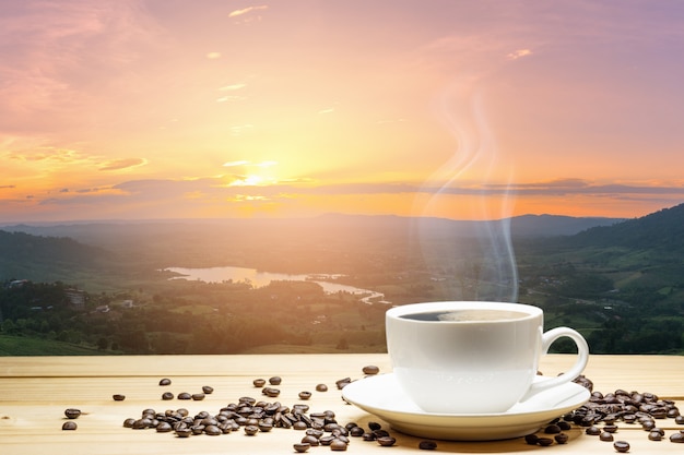 Taza blanca de café y granos de café en la tabla de madera con el fondo de la puesta del sol natural