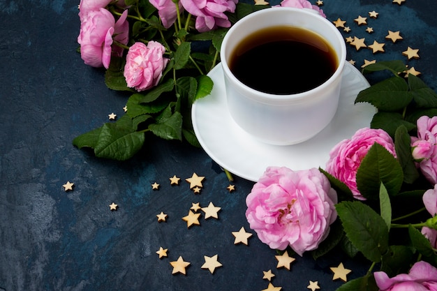 Taza blanca con café negro, estrellas y rosas rosadas sobre una superficie azul oscura