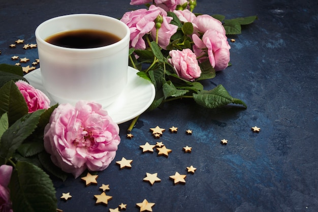 Taza blanca con café negro, estrellas y rosas rosadas sobre una superficie azul oscura