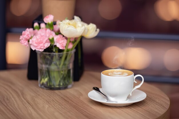 taza blanca de capuchino con arte y flores frescas en la mesa en la cafetería