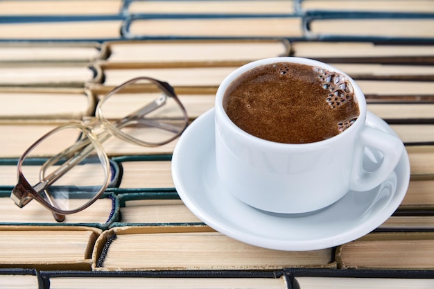 Una taza blanca de capuchino de café fragante y gafas de lectura contra el fondo de libros viejos gastados apilados boca abajo