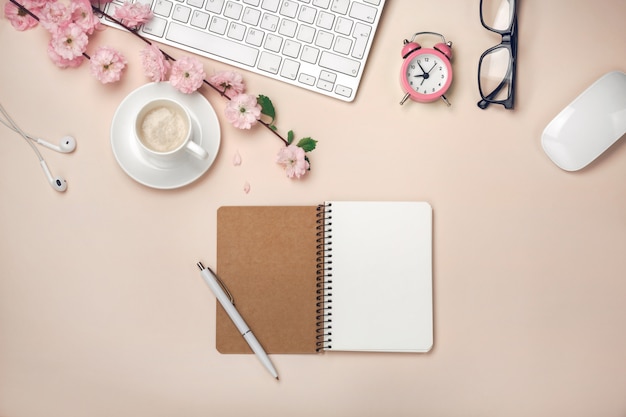 Taza blanca con capuchino, flores de sakura, teclado, reloj despertador, cuaderno sobre un fondo rosa pastel