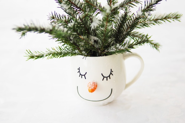 Taza blanca con la cara de un muñeco de nieve dormido y con ramas de abeto nevadas sobre un fondo blanco.