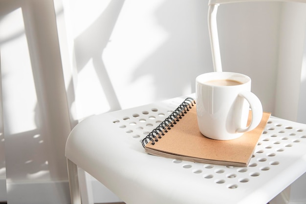 Taza blanca con cuaderno de café en una silla blanca bajo luz natural