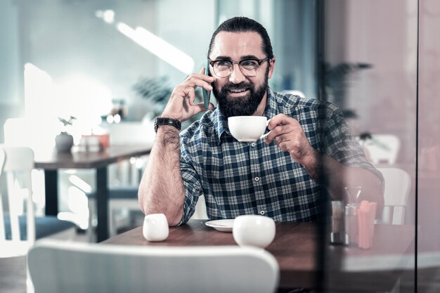 Taza blanca. Hombre maduro barbudo de ojos oscuros vistiendo camisa cuadrada sosteniendo una taza blanca de delicioso café