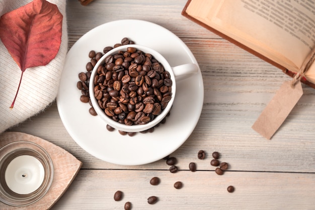 Una taza blanca llena de granos de café en una mesa de madera de color beige claro con un libro y un suéter. Vista superior con espacio para copiar.