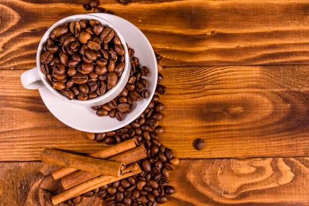 Taza blanca llena de granos de café y palillos de canela en una mesa de madera Vista superior