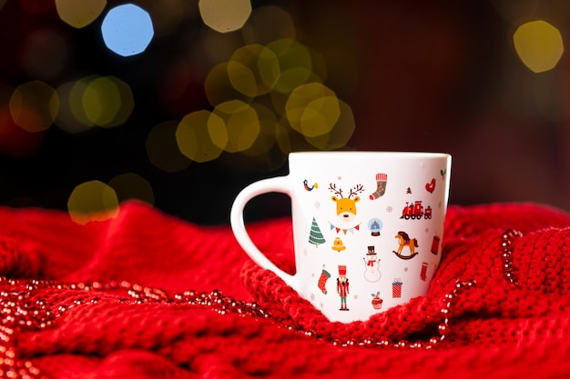 Taza blanca con un patrón de Año Nuevo sobre una manta de punto rojo sobre un fondo oscuro en el lateral, Bodegón navideño. Foto de alta calidad