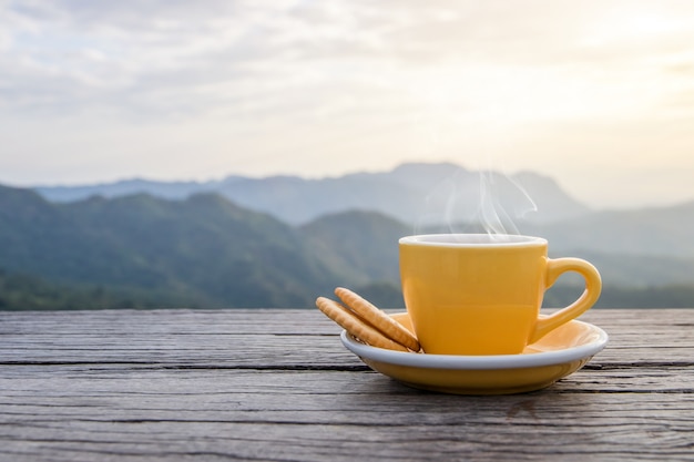 Una taza blanca de tazas de café exprés calientes colocadas con galletas en un piso de madera con niebla matutina y montañas con fondo de sol, mañana de café