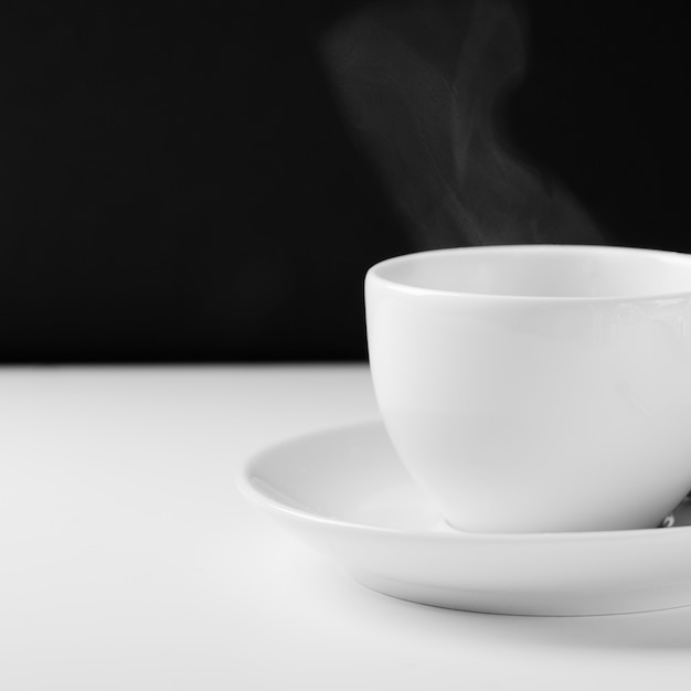 Taza blanca para el té con una bebida caliente en una tabla de madera blanca en un fondo negro.