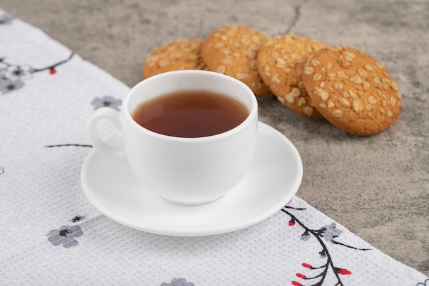 Una taza blanca de té caliente con galletas de avena sobre un mantel.