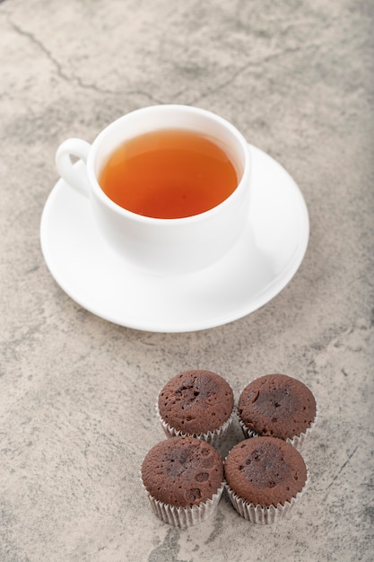Una taza blanca de té caliente con muffins de chocolate colocada sobre una mesa de piedra.