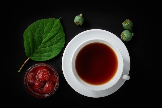 Taza blanca con té, mermelada de fresa, cajas de amapolas y hojas de hortensias en el espacio oscuro, vista superior, naturaleza muerta