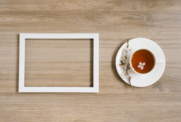 Una taza blanca con té negro y una ramita de flor de manzano y un marco blanco sobre un fondo de madera. Mocap y hasta prostranstva