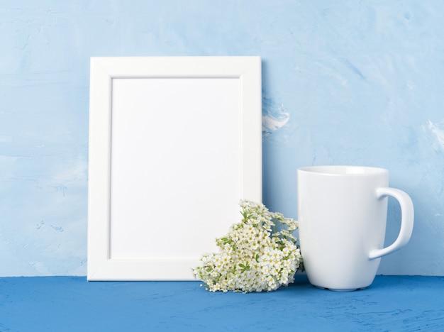 Taza blanca de té o café, marco, ramo de flores en la mesa azul frente a muro de hormigón azul
