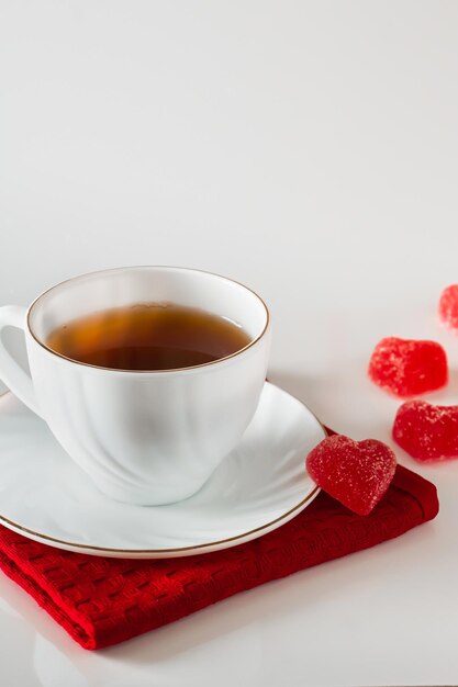 Taza blanca con té en una servilleta roja y mermelada en forma de corazón sobre un fondo blanco