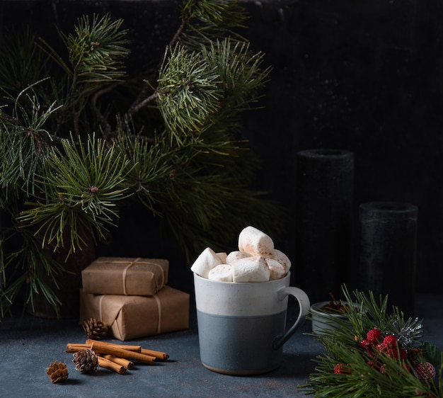 Taza con cacao, malvavisco sobre fondo azul oscuro con canela, velas, regalos y abeto. Imagen oscura y de humor