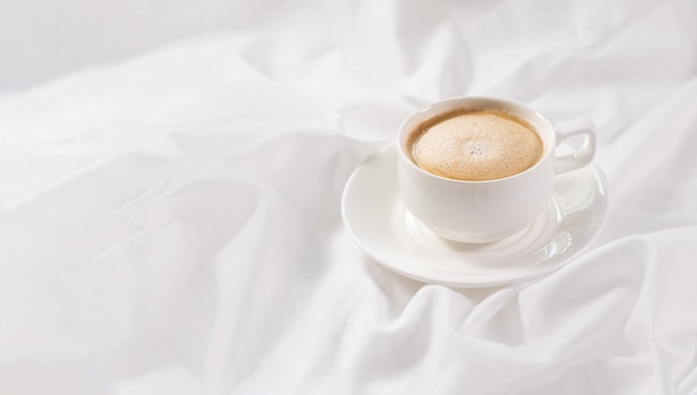 Foto taza de café сappuccino en la cama sobre una sábana blanca.