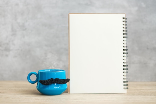 Taza de café azul o taza de té con decoración de bigote negro y bloc de notas de papel vacío o calendario en la mesa. Espacio de copia en blanco para texto. Noviembre azul, feliz día del padre y concepto del día internacional de los hombres