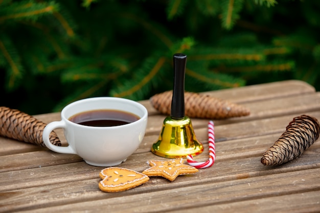 Taza de café, bastón de caramelo y campana con galletas en la mesa de madera con ramas de abeto en el fondo