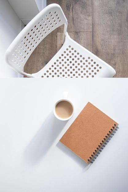 Taza de café blanca, un cuaderno sobre una mesa blanca, hay una silla al lado Vista superior plana
