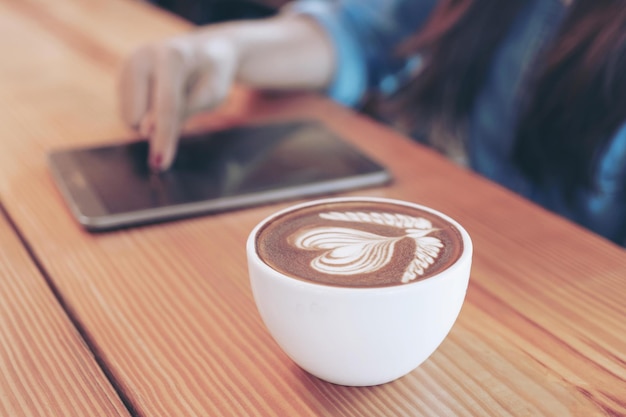 Taza de café blanca con patrón de corazón de arte latte sobre un fondo de mesa de madera y mano de mujer joven sosteniendo un teléfono inteligente en una cafetería