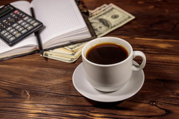 Taza de café y bloc de notas con dólares, lápiz y calculadora en escritorio de madera. concepto financiero