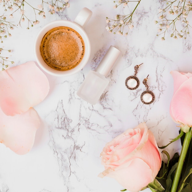 Foto taza de café; botella de barniz de uñas; rosas; pendientes y flores de aliento de bebé sobre fondo texturizado.