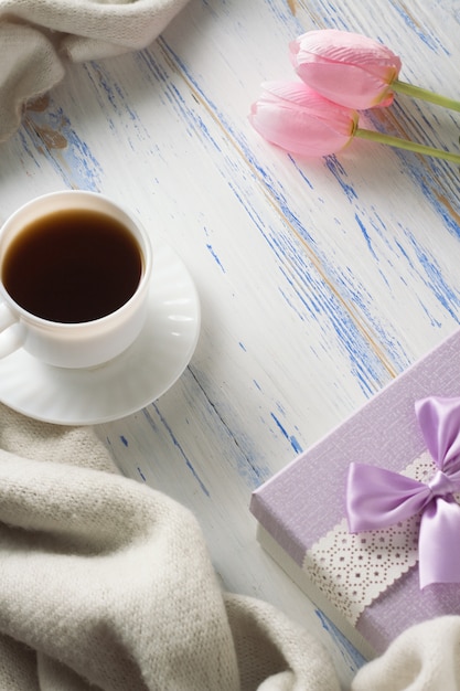 Taza con café, bufanda, regalo, tulipanes en la mesa de madera blanca. Concepto de primavera