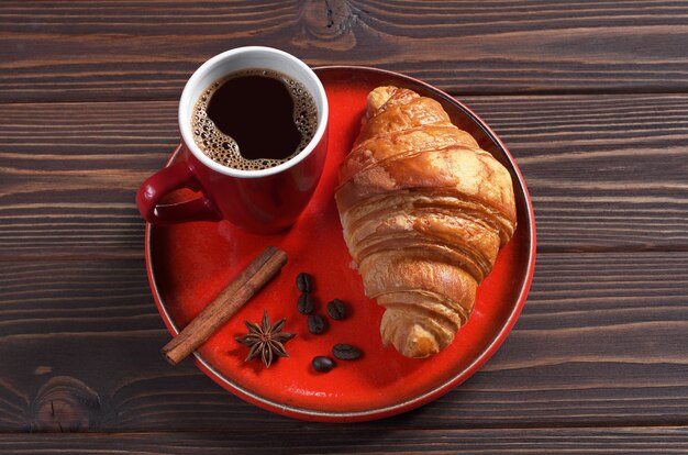 Taza de café caliente y croissant recién hecho en vajilla roja sobre mesa de madera oscura.