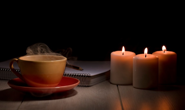 Taza de café caliente con cuaderno de humo amarillo con lápiz y velas encendidas Mesa de escritorio