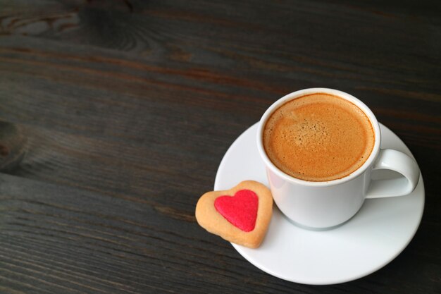 Taza de café caliente con una galleta en forma de corazón sobre fondo de madera negra