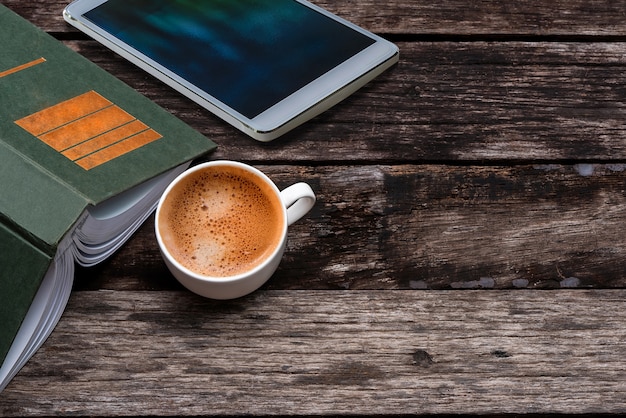 Taza de café caliente, libro verde y tableta blanca sobre fondo de mesa de madera