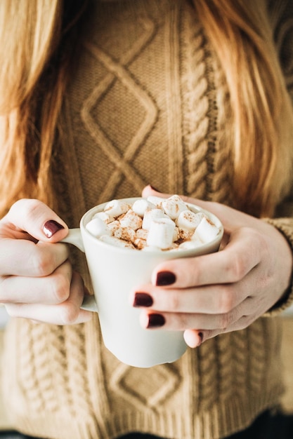 Taza con café caliente con malvavisco de crema batida y canela en manos femeninas