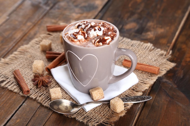Foto taza de café caliente con malvavisco sobre tela de arpillera con terrones de azúcar de canela y anís estrellado sobre fondo rústico de tablones de madera